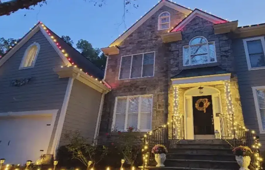 Gray stone house at dusk with red lights on the peaks and a glowing front entry.